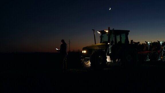 SLOMO晚上在田里在拖拉机旁边使用智能手机的农民
