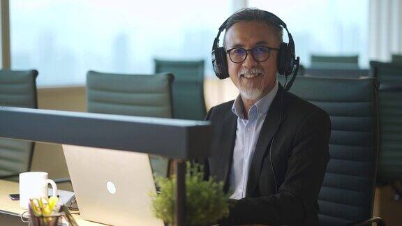 留着胡子的亚洲华人成熟商人戴着耳机看着工作站的摄像头