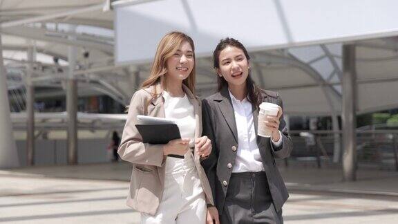 两个亚洲女商人拿着咖啡杯走在现代办公大楼的户外:慢动作