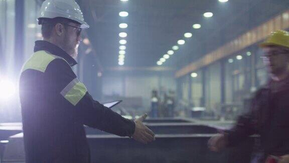 戴安全帽的工程师正带着一台平板电脑穿过一家重工业工厂是用握手来问候一个工人