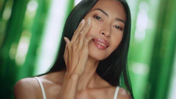 美丽的亚洲女人肖像轻轻应用面霜年轻的成年女性使用科学先进的天然化妆品护肤品使她的皮肤柔软光滑绿色森林背景