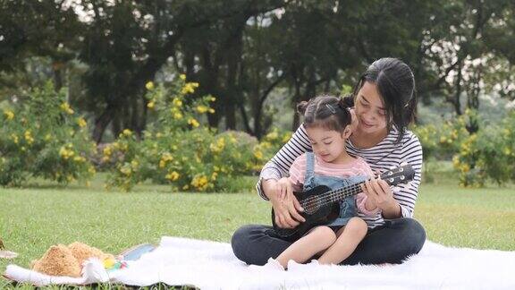 亚洲女人玩尤克里里与她可爱的女儿在公园野餐