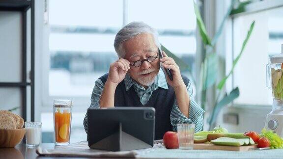 一名退休的亚洲男性头发灰白穿着休闲的衣服在现代公寓的餐桌上悠闲地吃着早餐喝着健康的饮料阅读平板电脑上的新闻和朋友聊天