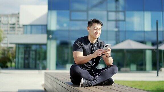 一名成年亚洲男子戴着耳机坐在一座现代城市大楼前的长凳上听音乐穿着运动服的英俊男子拿着智能手机
