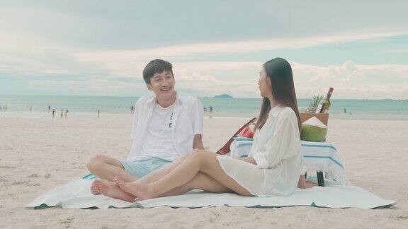 亚洲的年轻夫妇坐在野餐毯上在白色的沙滩和海边放松热带水果作为背景夏季、假期、假期和快乐的泰国人概念
