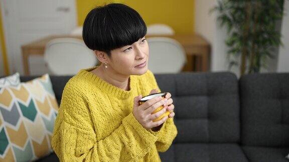 年轻的中国女子坐在家里的沙发上喝咖啡