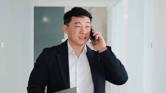 中国英俊的商人站在办公室大厅里面带愉快的微笑正在讨论打电话成功自信的商人有友好的谈话拿着笔记本电脑