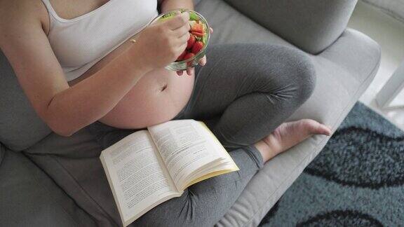 孕妇吃着简单的早餐早上在家里的沙发上读着一本书