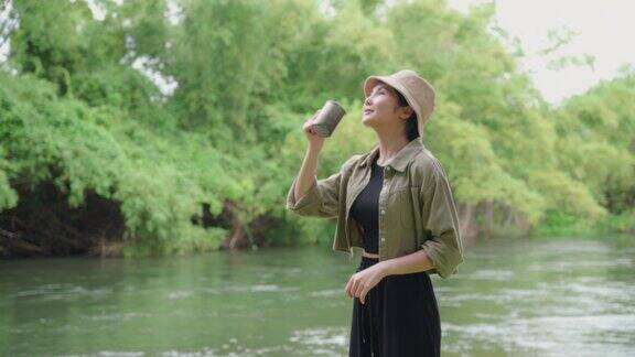 快乐的亚洲女人早上站在河边喝咖啡在大自然中露营她感到放松和清爽休闲旅行和户外活动的生活方式