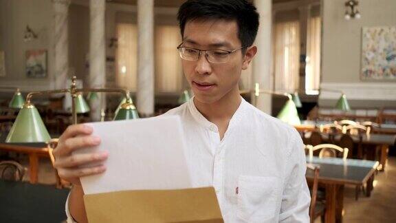 年轻的亚洲男学生在大学图书馆激动地打开装有考试成绩的信封如释重负地叹息