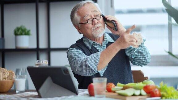 一名退休的亚洲男性头发灰白穿着休闲的衣服在现代公寓的餐桌上悠闲地吃着早餐喝着健康的饮料阅读平板电脑上的新闻和朋友聊天