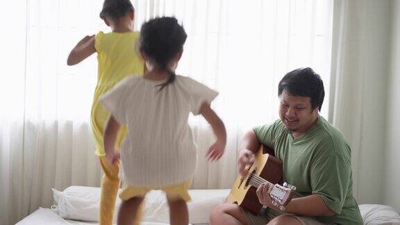 快乐的父亲和两个女儿在家里一起玩耍父亲在弹吉他女儿们在床上又跳又跳