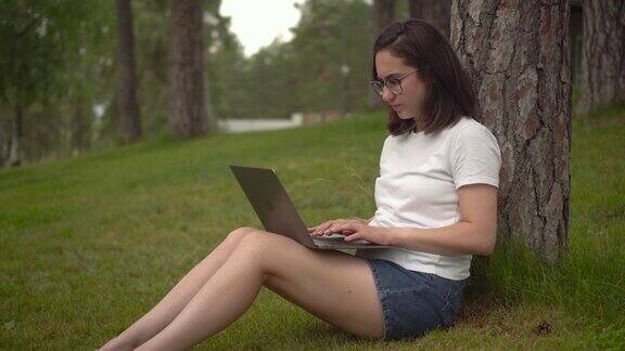 在大自然中拿着笔记本电脑的年轻女子一个戴眼镜的女孩坐在树下用笔记本电脑打字自由职业者的女人特写镜头4k