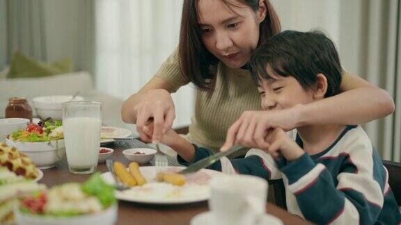 亚洲美丽的母亲帮助她的小可爱6岁的儿子吃鸡棒用叉子和刀在家里的餐厅早餐和家人一起享用美味的晚餐