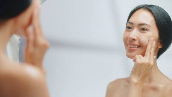 美丽的亚洲妇女肖像轻轻应用面霜面膜感性的触摸看在浴室的镜子女性用天然化妆品和护肤品使她的皮肤柔软慢动作