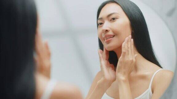 美丽的亚洲女人她完美柔软的脸皮肤使用先进的有机霜微笑在镜子里快乐的女性享受她的早晨例行公事采购产品健康自然美容化妆品