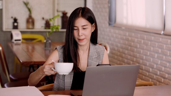 漂亮的亚洲女孩工作日举起一杯白咖啡喝拿铁在面包店用笔记本电脑工作时用手指按手机与朋友聊天
