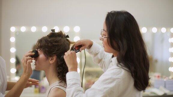 女发型师在美容工作室用卷发钳卷年轻女子的头发化妆师用海绵在女士脸上涂抹粉底