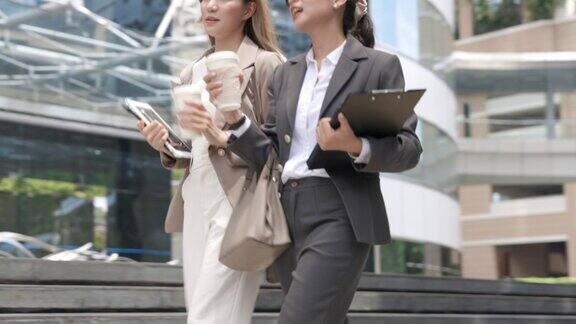 两个亚洲女商人拿着咖啡杯走在现代办公大楼的户外