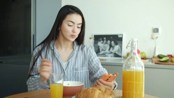 快乐的亚洲女性在家里的厨房里吃着羊角面包喝着果汁用着智能手机