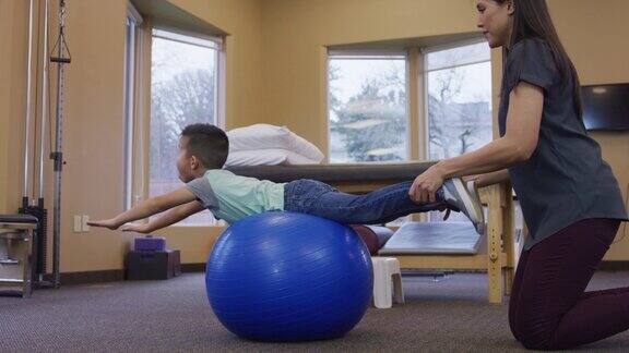 小男孩躺在一个运动球上而他的理疗师按住他的腿