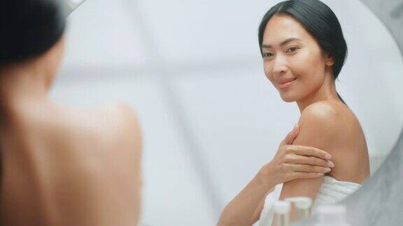 美丽的亚洲女人在她完美的肩膀上应用身体霜使皮肤柔软在镜子里感觉微笑快乐的女性享受她的美丽健康天然化妆品护肤品
