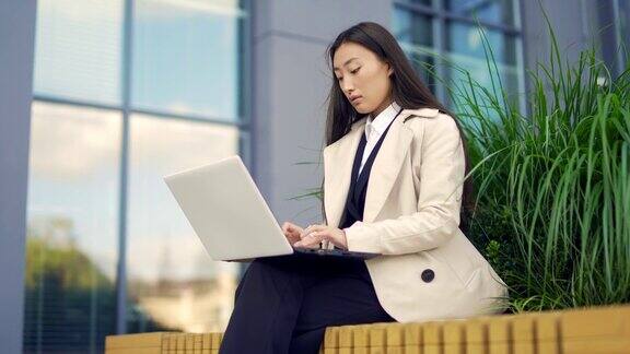 亚洲女性自由职业者在线工作使用打字笔记本电脑坐在长凳外的现代办公大楼在城市公园的街道
