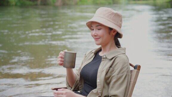 快乐的亚洲女人早上坐在河边喝咖啡在大自然中露营她感到放松和清爽休闲旅行和户外活动的生活方式