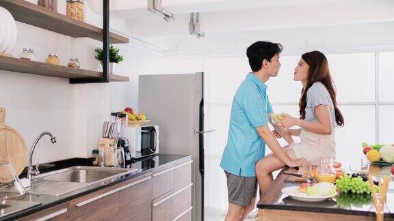 快乐的亚洲情侣以一种有趣而浪漫的方式聊天、准备和烹饪当女性坐在家里的厨房柜台上一起在家里的厨房里喂薯片时4k视频实时播放