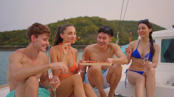 一群不同的朋友在游艇上吃烧烤迷人的年轻男女聚在一起庆祝节日双体船在夏日夕阳下航行