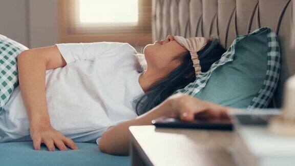 年轻的亚洲女性穿着睡衣戴着睡眠面罩躺在方便和舒适的床上睡眼惺忪地醒来感到手机嘈杂的闹铃早上晚些时候在家里的卧室叫醒她