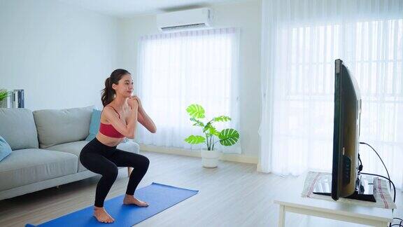 亚洲美女呆在家里在家做有氧运动迷人的女孩正在做禁闭活动蹲下锻炼遵循在线健身教练的指导视频