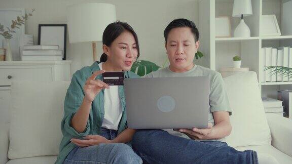 幸福的亚洲夫妇一起坐在家里客厅的沙发上用笔记本电脑购物用信用卡在线支付生活方式花时间在家里