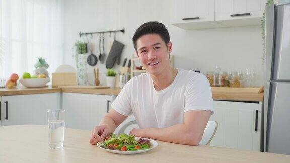 亚洲活跃的帅哥拒绝吃甜甜圈喜欢吃健康食品年轻有魅力的男性在厨房的碗里吃蔬菜沙拉在桌子上喝干净的水饮食与健康理念