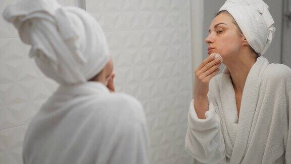 漂亮的女士站在明亮的浴室里用棉垫照镜子护肤卸妆净化乳液或胶束水