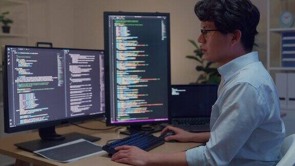 年轻的亚洲男性软件开发人员使用电脑编写代码坐在有多个屏幕的桌子上晚上在办公室工作程序员开发