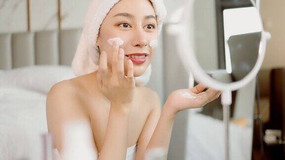 一段迷人年轻女子在家浴室里的4k视频美丽的年轻亚洲女性愉快地应用霜或乳液与保湿面美丽的亚洲女孩应用护肤触摸面部化妆健康和健康的conc