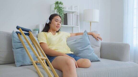 亚洲年轻截肢女子在家里的客厅听音乐迷人的没有腿的女性感到快乐和放松坐在沙发上戴着耳机听着歌在家里度过闲暇时光