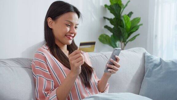 亚洲美女喜欢在家里的客厅网上购物漂亮的女孩坐在沙发上用手机在网上商店购买商品后拿着信用卡进行网上银行支付
