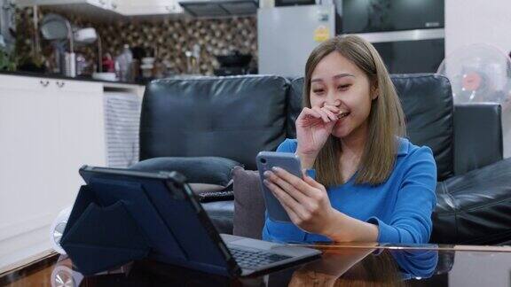 年轻的亚洲女性坐在家里使用手机和笔记本电脑工作