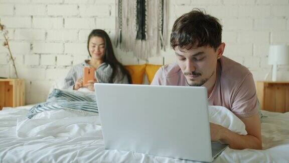 男人在床上用笔记本电脑工作而妻子在微笑着使用智能手机