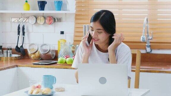 年轻美丽的亚洲女人谈论智能手机与幸福坐在厨房吃零食在家里人们在电话生活方式