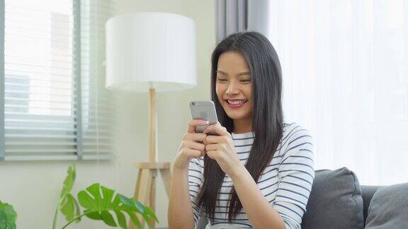 亚洲美女坐在家里的沙发上用手机聊天快乐迷人的年轻女孩在家里度过休闲时间感到放松享受在客厅用智能手机交流和讨论