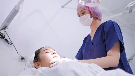 侧视图的医疗妇女的脸躺在白色的医疗沙发上医生用卫生手套发帽给医疗服务年轻的亚洲妇女调整手术灯到脸检查皮肤质量