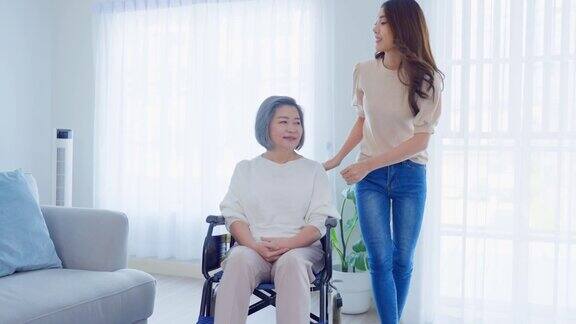 亚洲年轻的女儿在家照顾坐轮椅的残疾老太太美丽的女孩帮助和照顾年迈成熟的残疾母亲病人做物理治疗在客厅在家里