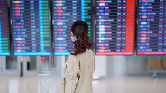 年轻的亚洲女子戴着口罩手持护照和登机牌在国际机场看着航班信息板查询她的航班