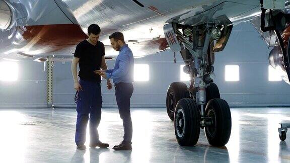 在机库飞机维修工程师向飞机技术员展示平板电脑上的技术数据他们站在干净的新飞机旁边