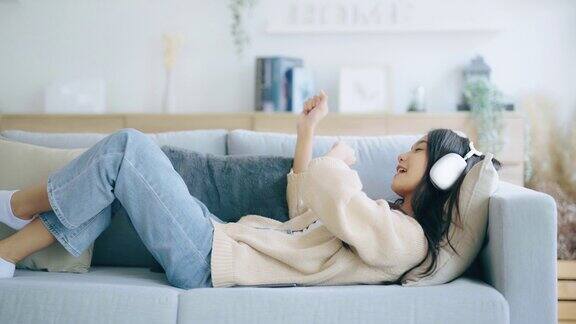 年轻的亚洲女子在家里客厅的沙发上听音乐快乐亚洲女性使用手机戴耳机躺在沙发上