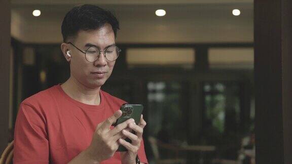 戴着眼镜的亚洲中年男子正在用智能手机和蓝牙耳机轻松愉快地观看娱乐电影通过手机输入通信信息