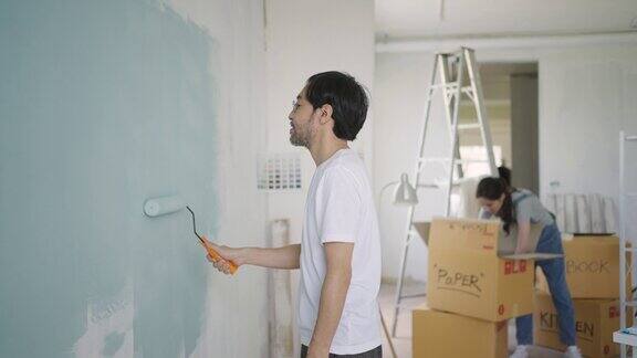 一对年轻的亚洲夫妇在装修他们的新公寓时正在用滚轴用蓝色油漆修补墙壁